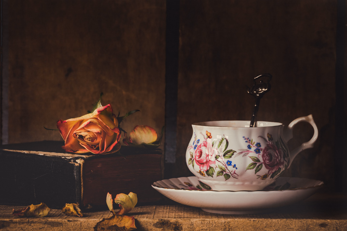 A floral porcelain teacup on its saucer with an ornate spoon, beside an old leather book and a faded orange rose on a dark wooden shelf