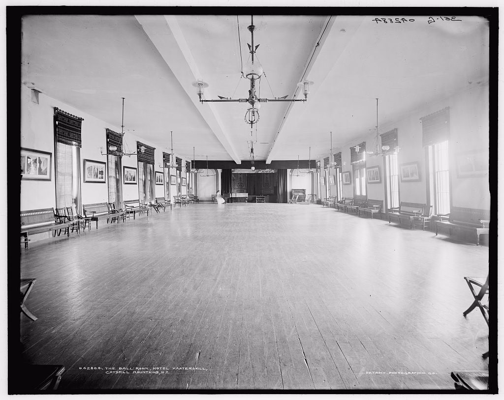 Black and white photograph of a long hotel ballroom with a bare polished wood floor and one figure seated near the stage, wooden benches and chairs lined up against both walls under framed pictures, hanging light fixtures, and a curtained stage at the far end