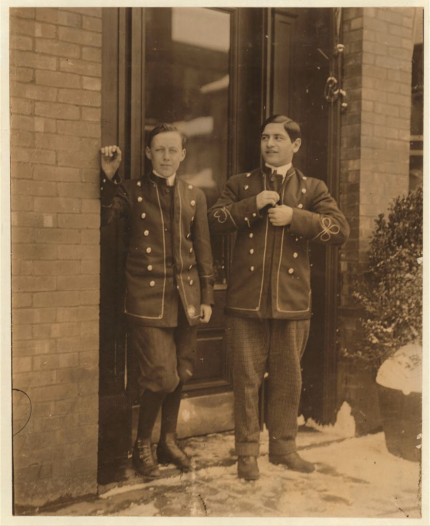 Sepia photograph of two young bellboys standing at a hotel doorway on a snowy sidewalk, both in short fitted jackets with stand collars, light piping down the fronts and two rows of round buttons, one in knee breeches and dark stockings and the other in long checked trousers