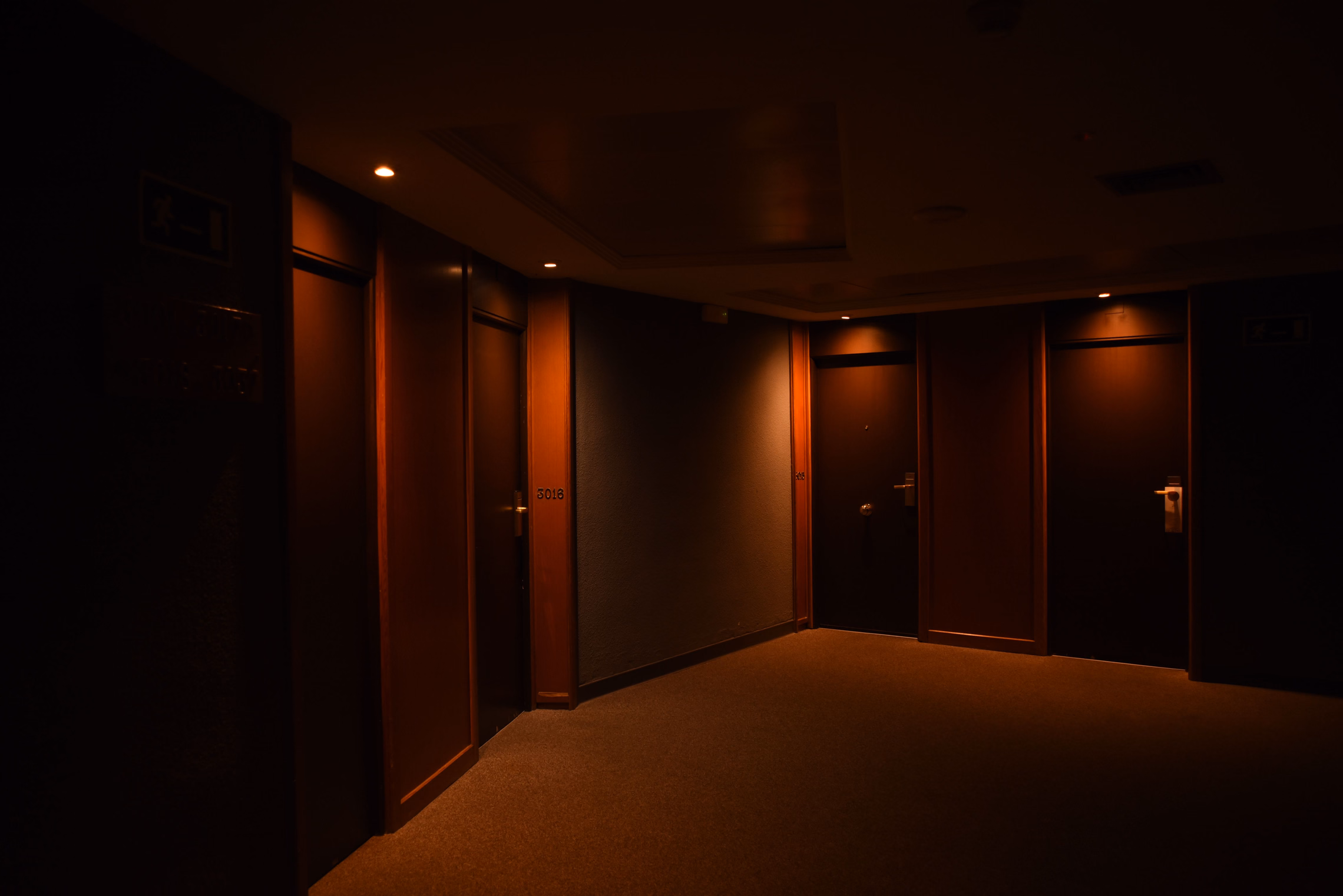 A dark hotel corridor with closed wooden doors and a carpeted floor, lit only by a few warm orange ceiling lights