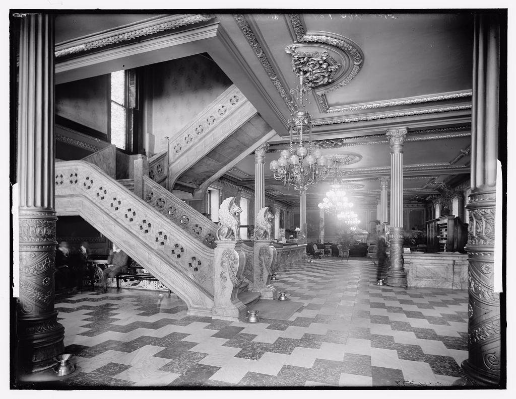 Black and white photograph of a hotel office and foyer with a marble staircase whose newel posts are carved griffins, fluted columns, crystal chandeliers and a checkerboard marble floor running back to the desk