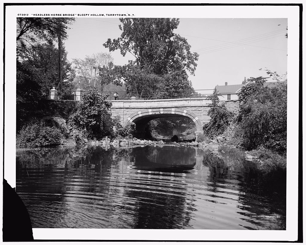 Black and white photograph of a stone arch bridge over a still stream in Sleepy Hollow, trees on both banks and a man standing on the bridge, with the negative's caption Headless Horse Bridge, Sleepy Hollow, Tarrytown, N.Y.
