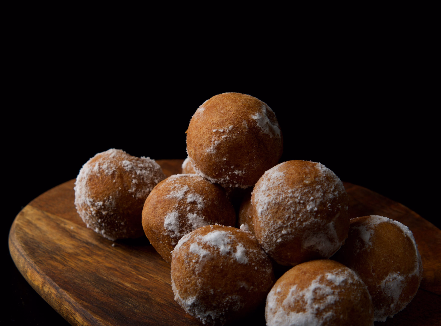 A pile of round, sugar-dusted fried dough balls on an oval wooden board against a black background