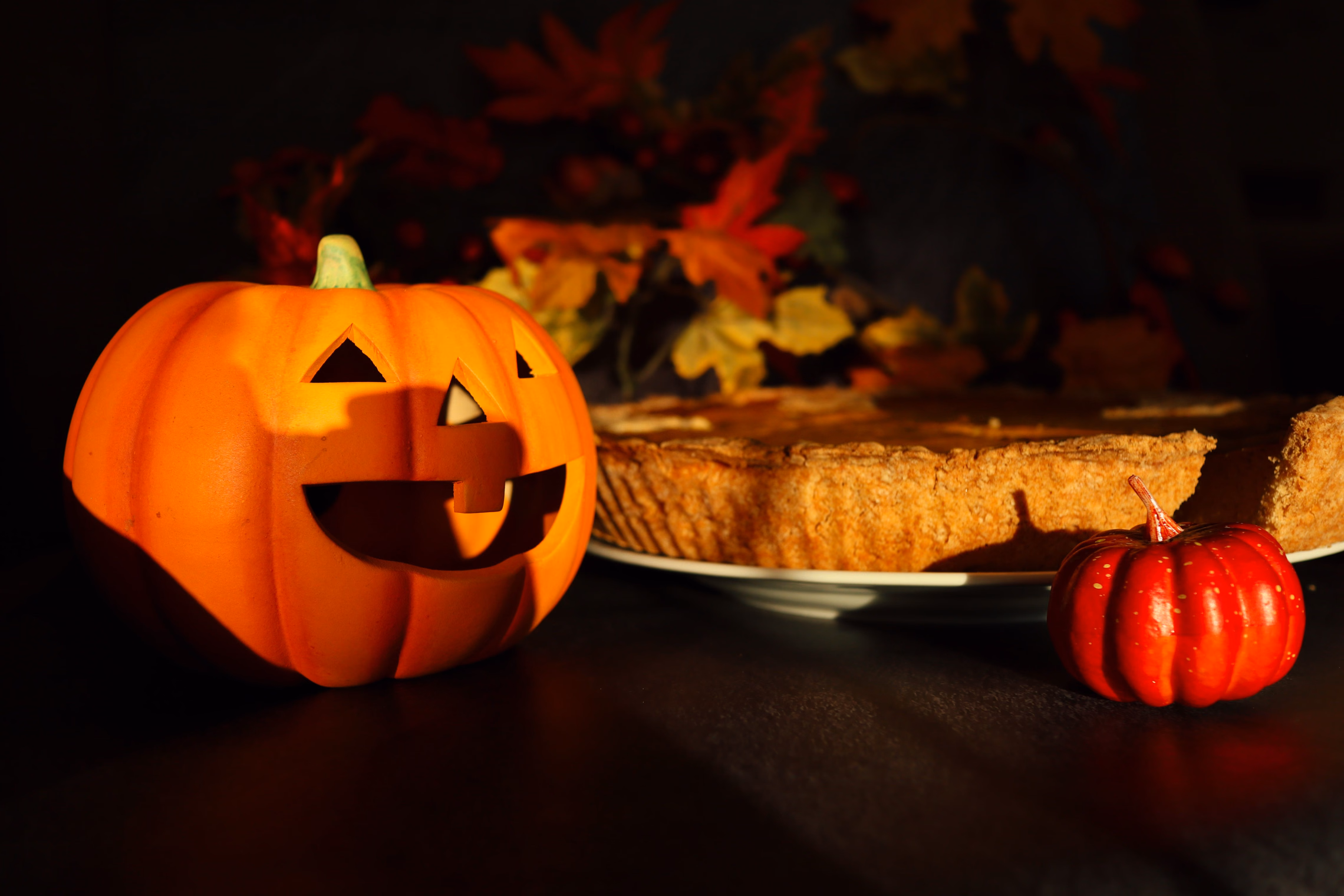 A jack-o'-lantern with triangle eyes beside a pumpkin pie on a plate and a small red pumpkin, lit by low sun against orange and red autumn leaves on a dark background