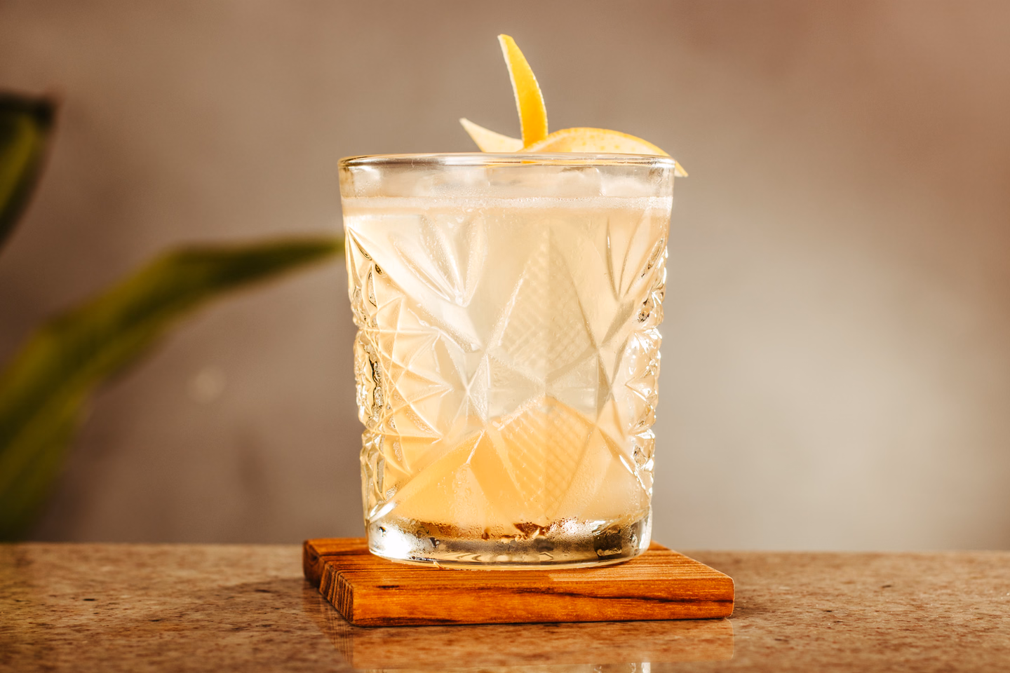 A pale, cloudy citrus cocktail in a cut-crystal glass with a curl of lemon peel, on a small wooden coaster