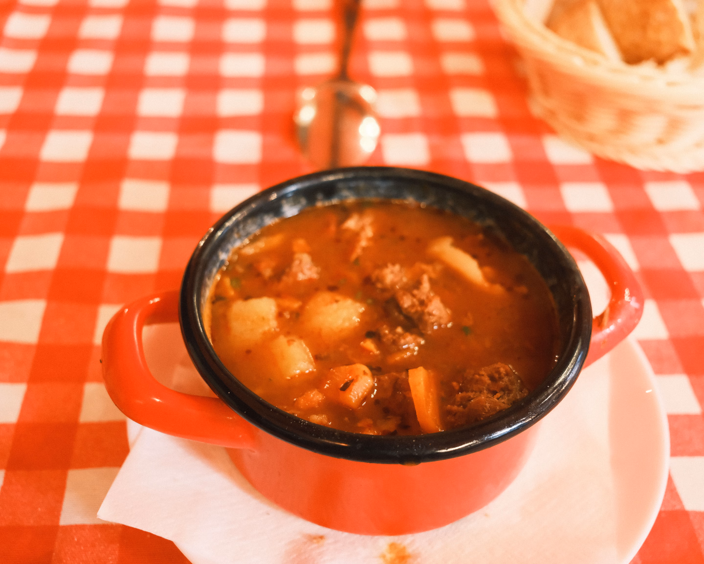 A red enamel pot of goulash with chunks of beef and potato on a red and white checked tablecloth, a basket of bread behind it