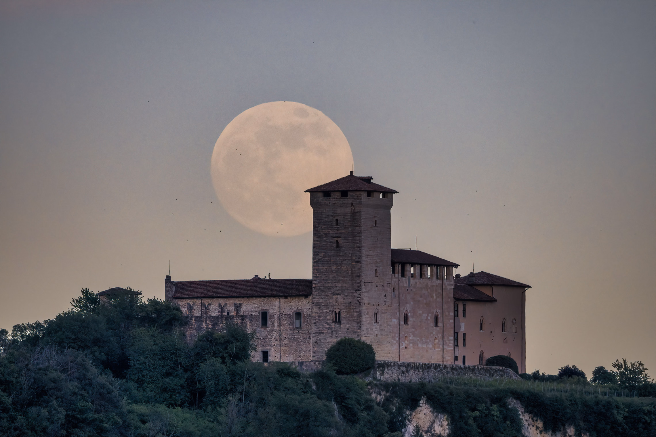 A full moon rising behind the square stone watchtower of a hilltop castle at dusk, with dark trees along the slope below