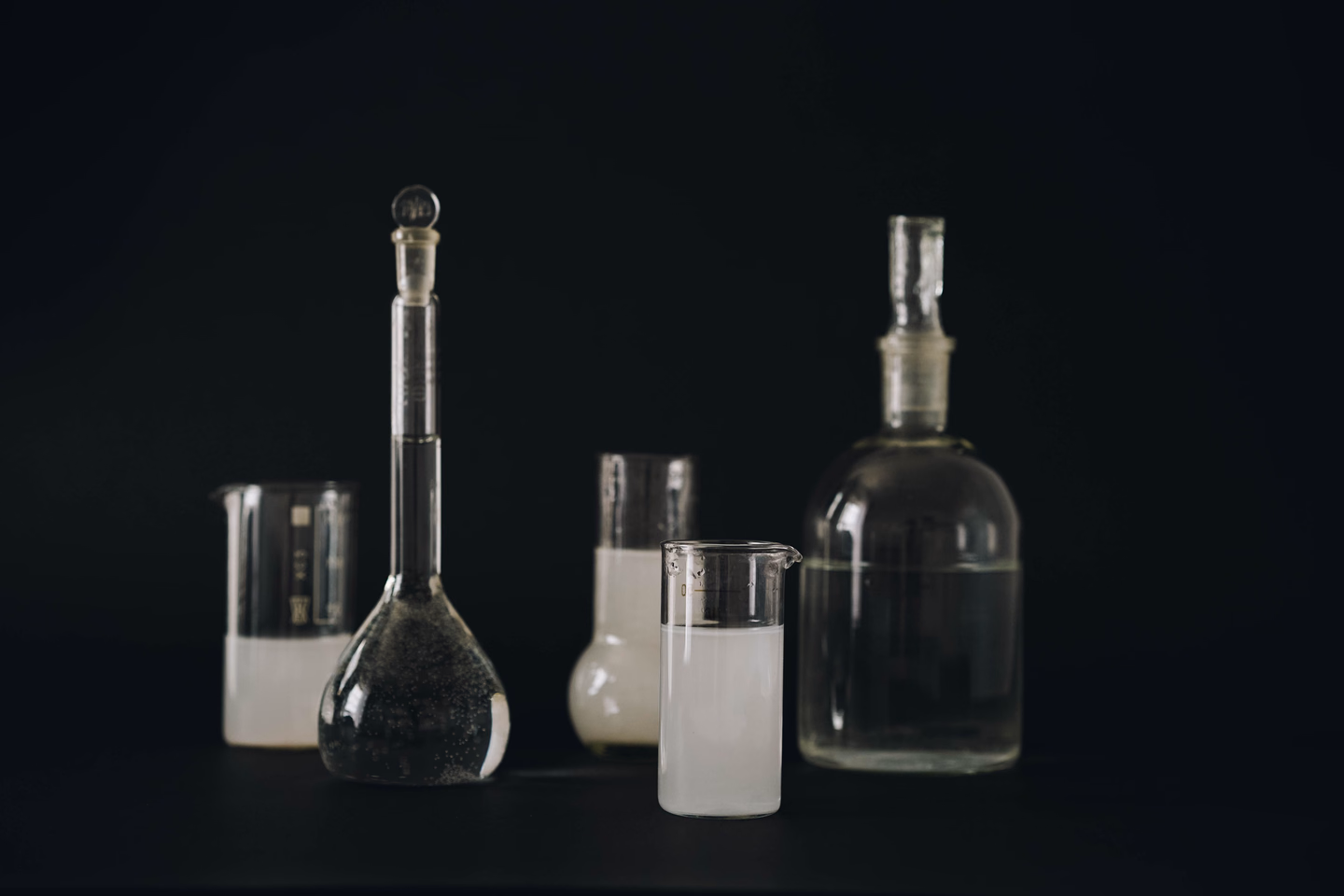 Laboratory glassware on a black surface: a tall volumetric flask, beakers and a stoppered bottle, some holding cloudy white liquid
