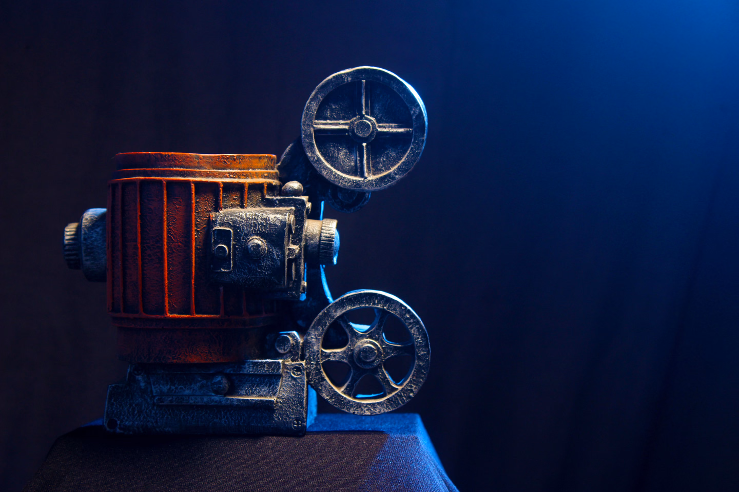 An old metal film projector with two reel arms, lit from the side against a dark blue curtain