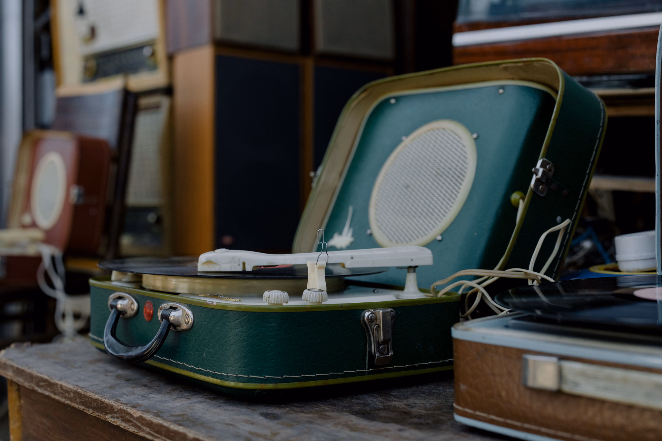 A green suitcase record player with its lid open and a black record on the platter, on a worn wooden table in front of old radios and speakers