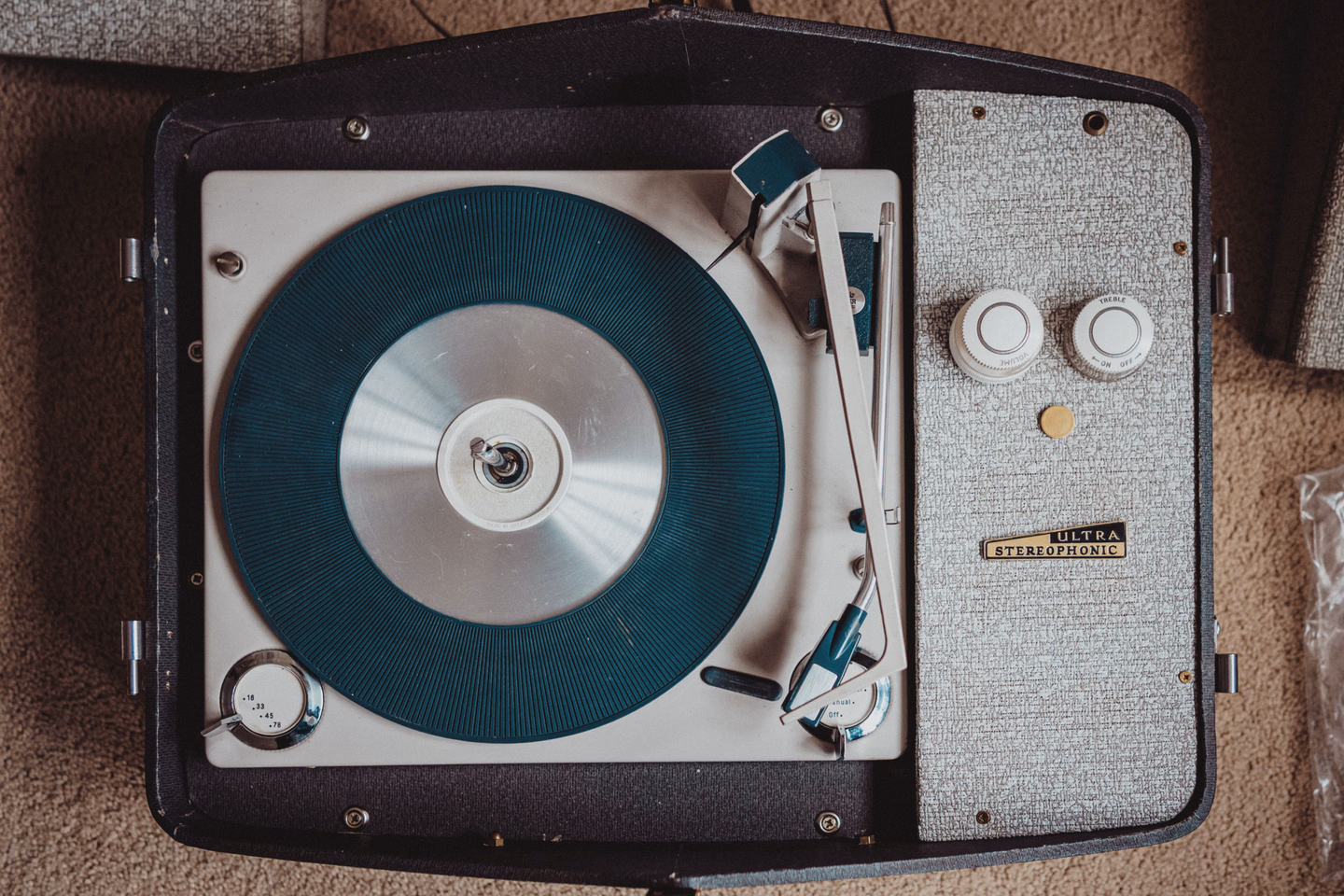 Top view of an open portable record player in a black case with a gray speckled speaker panel, a teal ribbed platter mat, a speed selector marked 16, 33, 45 and 78, and a badge reading Ultra Stereophonic