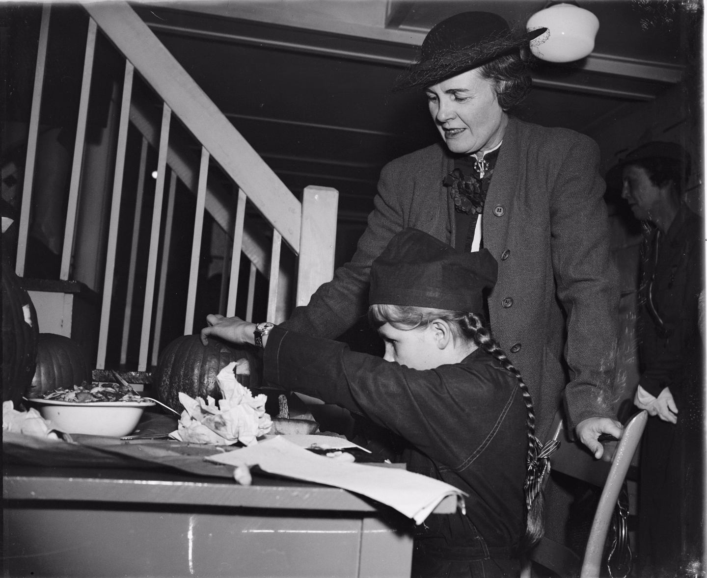A woman in a hat and coat guides a Girl Scout in uniform and braids as she cuts into a pumpkin at a table