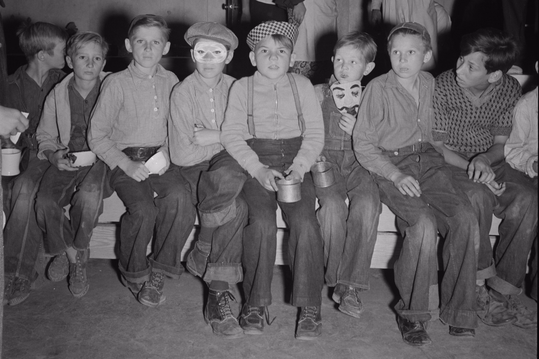 A row of boys in caps and denim sitting on a bench at a 1938 Halloween party, one wearing a paper eye mask and another holding a mustached false face