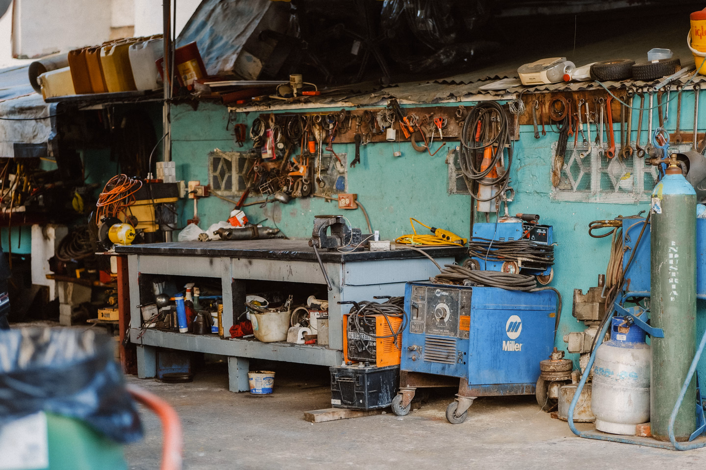 A cluttered garage workshop with a workbench, welding machines, hanging tools and gas cylinders standing on the concrete floor