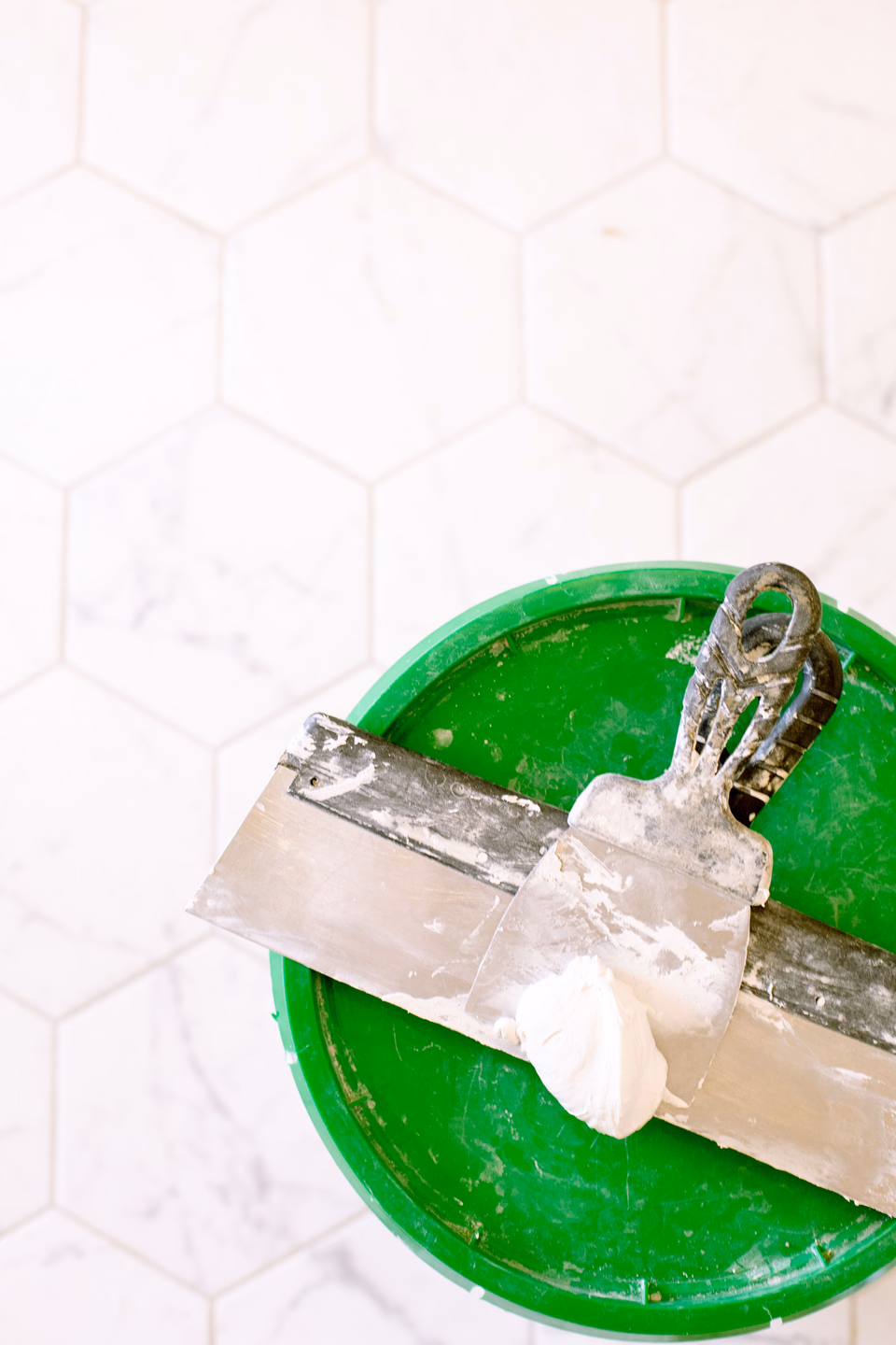 Overhead view of a green bucket lid with two taping knives resting across it, a dollop of white joint compound on the smaller blade, on white hexagon tile