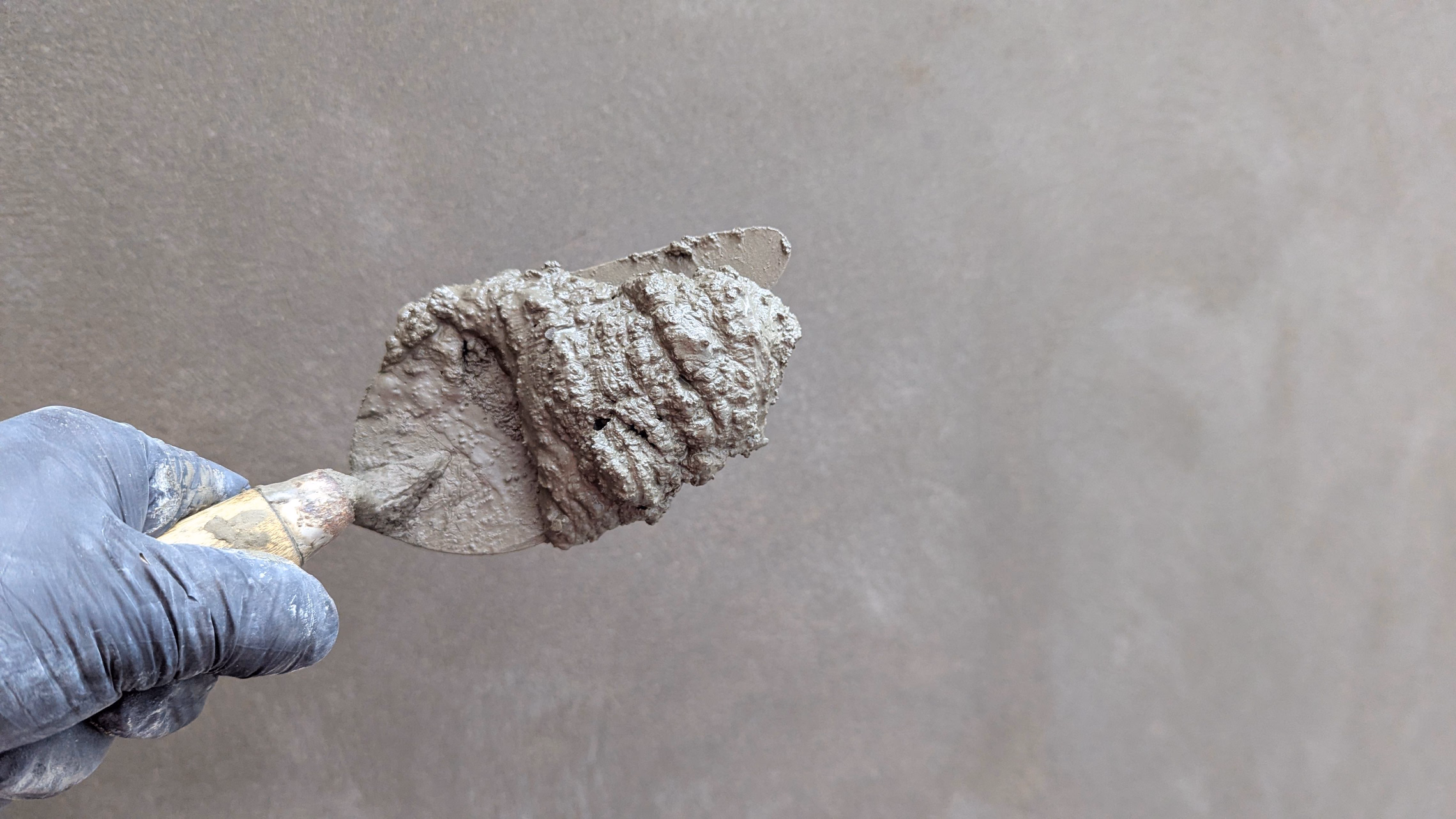 A gloved hand holding a trowel loaded with wet gray plaster against a smoothly plastered gray wall
