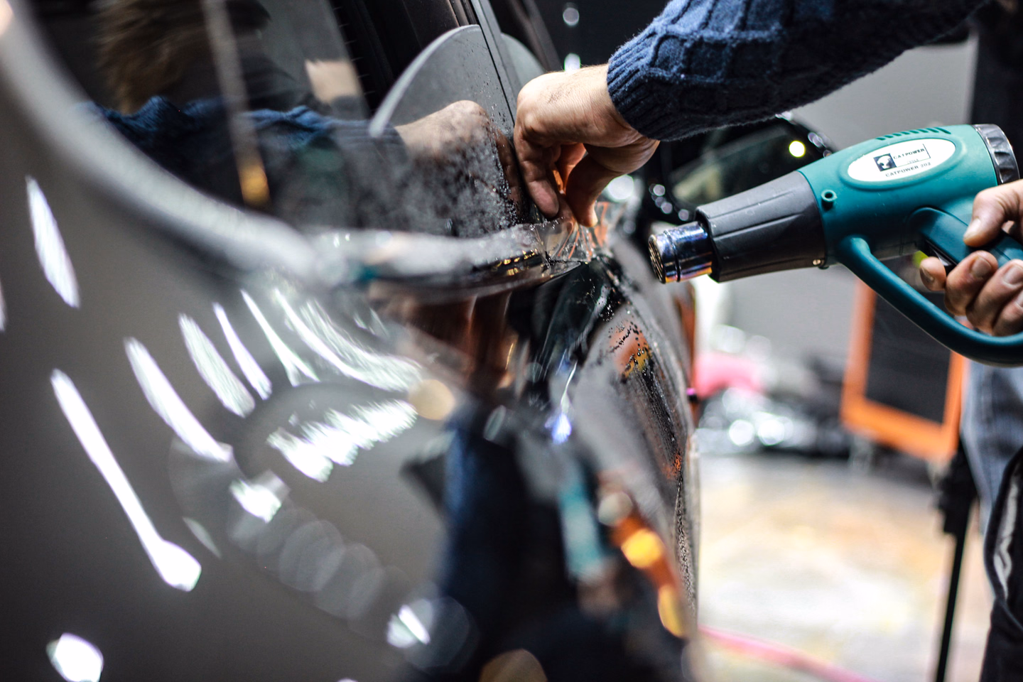 A technician's hand smoothing tinted film onto a car window while the other hand aims a teal heat gun at it