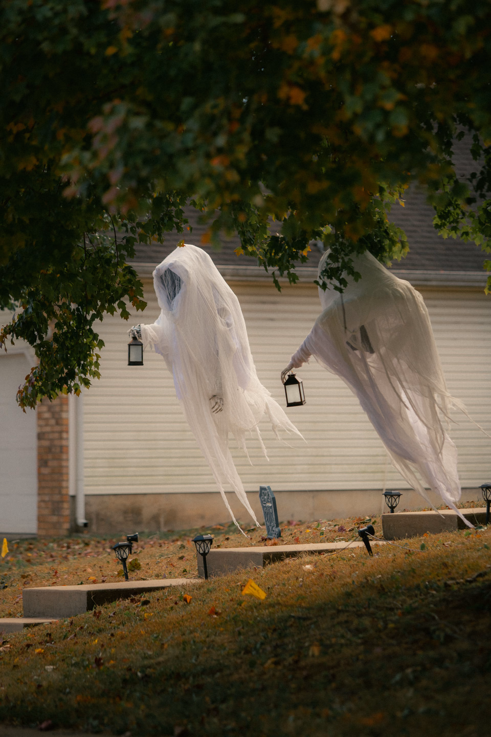 Two cheesecloth ghosts carrying small lanterns hang from tree branches above a sloping front yard with path lights