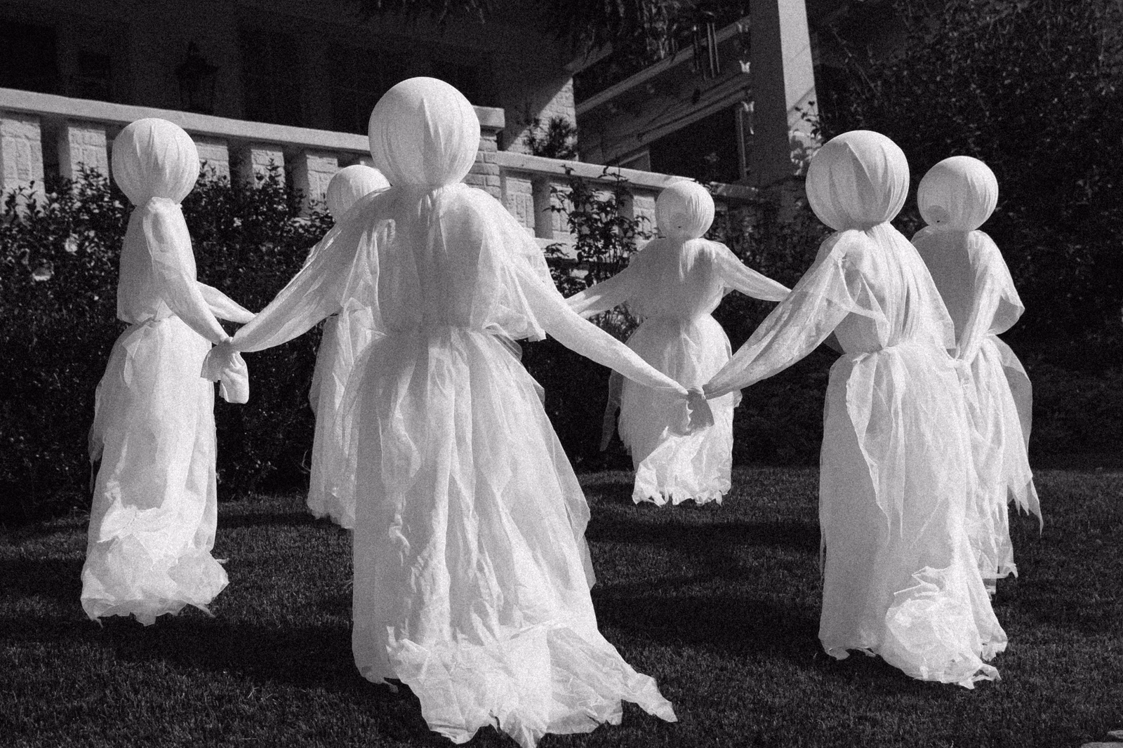 Black and white photo of sheer white cloth ghosts with round heads holding hands in a circle on a front lawn