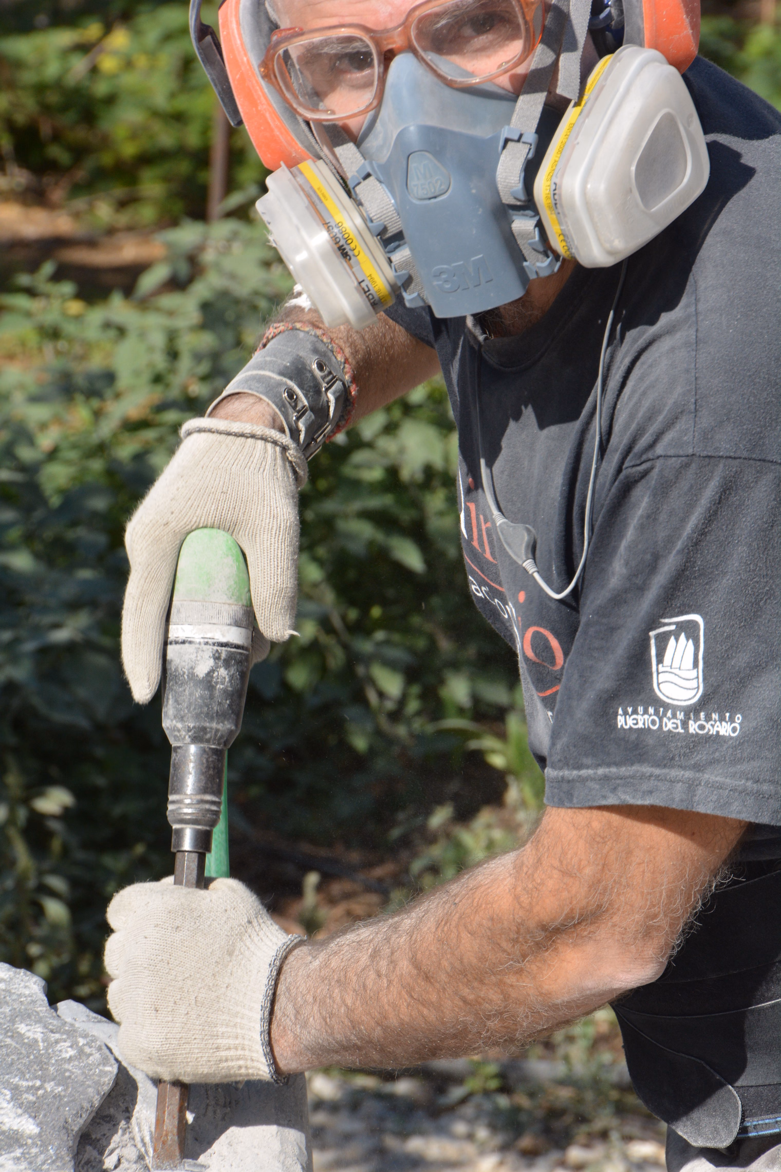 A stone carver in a half-face respirator with twin cartridges, safety goggles and work gloves guiding a pneumatic chisel into a block of stone outdoors