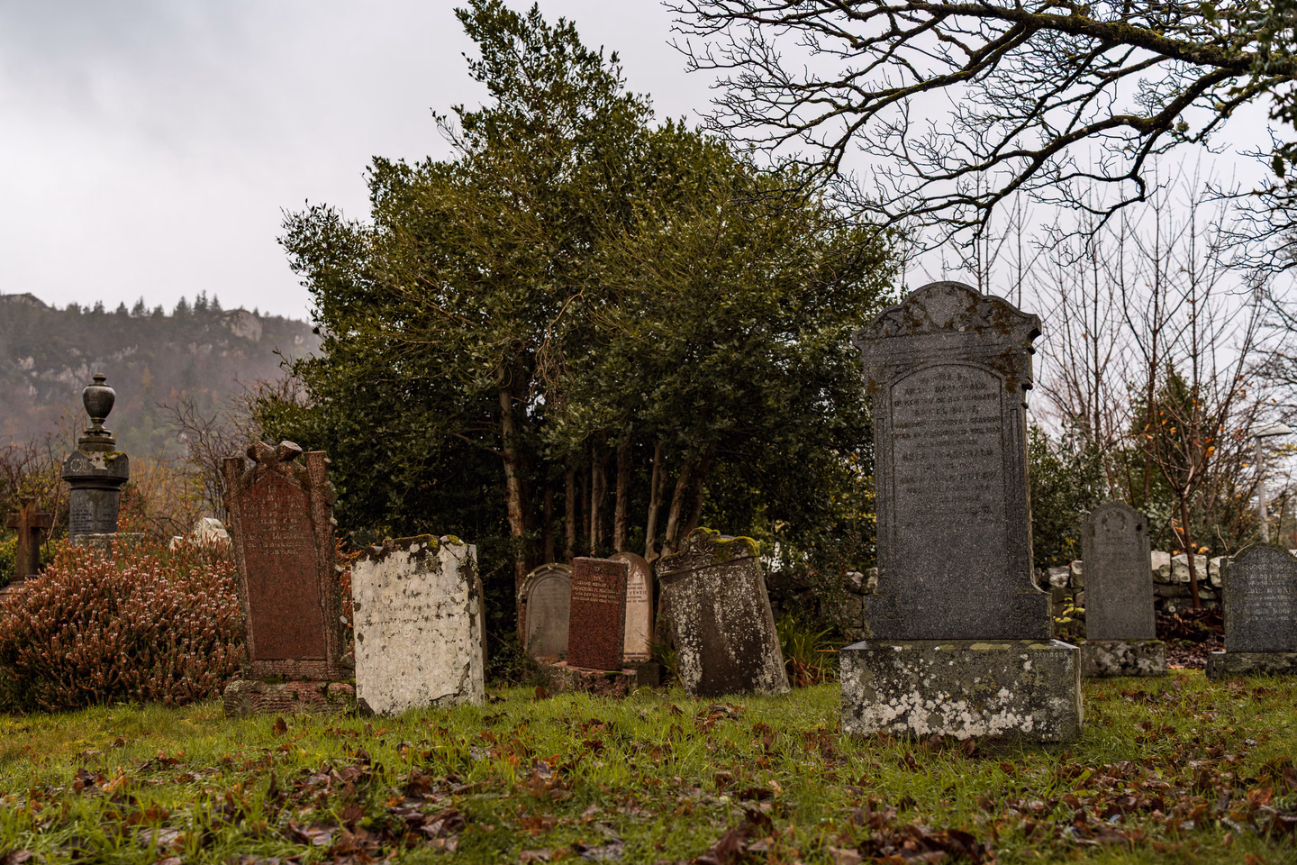 An old cemetery of mismatched headstones in rough grass and fallen leaves, some pale and stained, some dark red stone, a few tilted, with a tall gray monument on a stepped base at the right